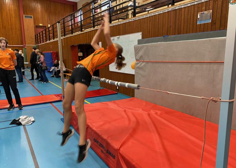 En kvinnlig idrottare i orange tröja försöker hoppa höjdhopp i en inomhusgymnastiksal, medan andra tittar på i bakgrunden.