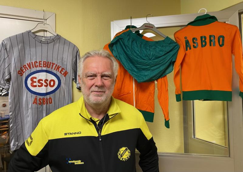 En äldre man i gul och svart sportjacka står inomhus. Bakom honom hänger gamla sporttröjor och jackor, en med