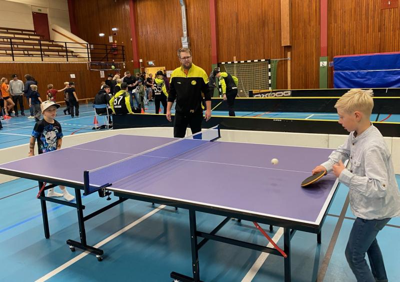 En pojke spelar bordtennis med en vuxen i ett gym medan ett annat barn och flera personer tittar på i bakgrunden.