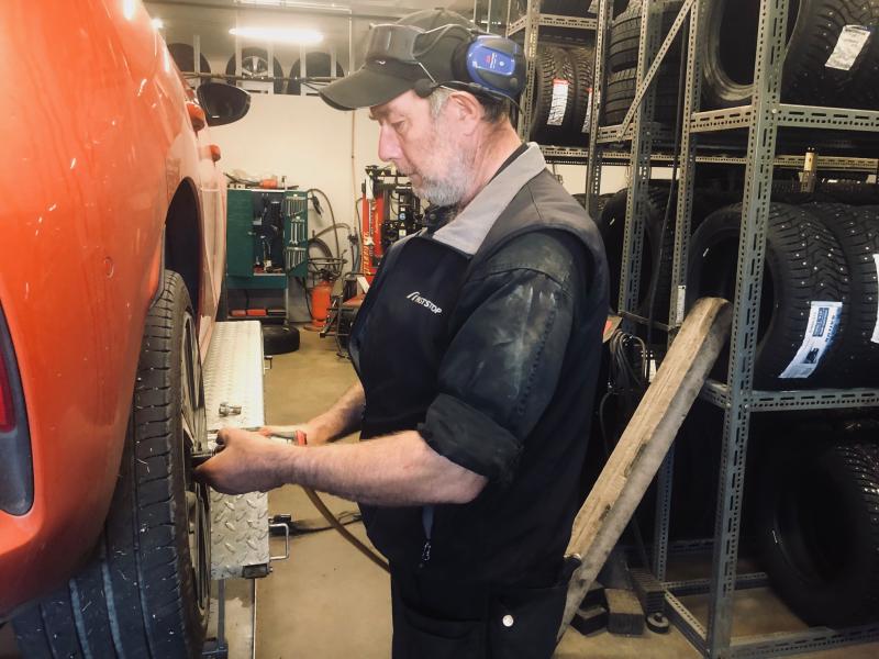 En mekaniker med skyddshörlurar använder ett verktyg för att arbeta på hjulet på en röd bil i en bilverkstad med däck på hyllor i bakgrunden.