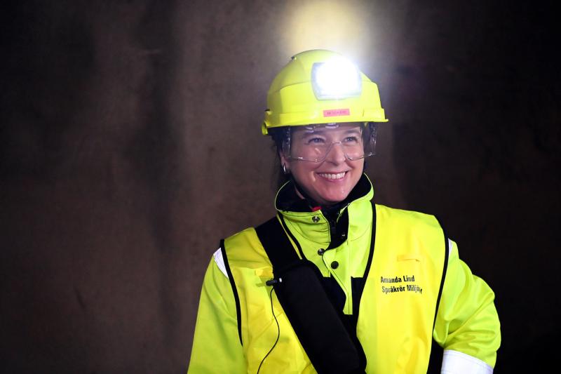En person som bär en gul skyddshjälm med pannlampa, klara skyddsglasögon och en varseljacka står och ler mot en mörk bakgrund.