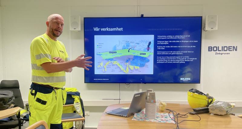 En man i varselkläder presenterar sig i ett mötesrum och gestikulerar mot en skärm som visar en karta och svensk text om Bolidens verksamhet. Skyddsutrustning ligger på bordet.