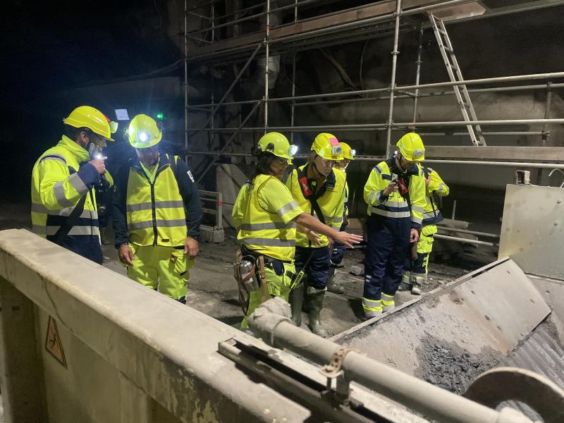 Fem arbetare i varselkläder och hjälmar inspekterar industriutrustning i en svagt upplyst lokal med metallställningar i bakgrunden.