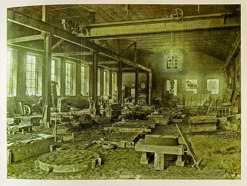 Svartvitt foto visar en gammal industriverkstad med stora fönster, maskiner, utspridda verktyg och konstruktionsbalkar under ett trätak.