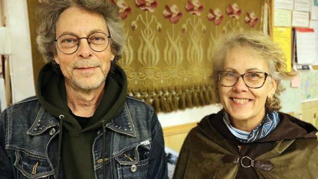 Två äldre personer med glasögon, den ena i jeansjacka och huvtröja, den andra i brun mantel, står inomhus framför en dekorativ gobeläng och en vägg med papper.