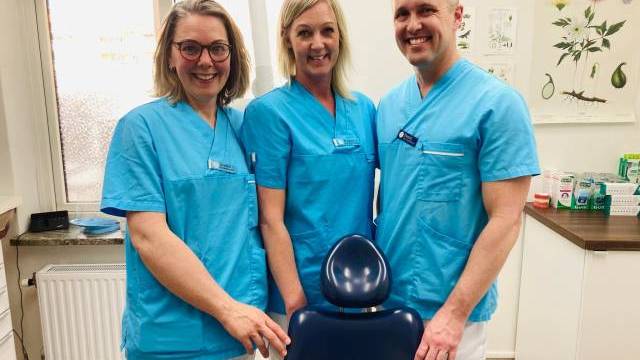 Tre tandläkare i blå scrubs leende på en tandläkare, stående bakom en tandläkarstol med tandvårdsutrustning synlig i bakgrunden.