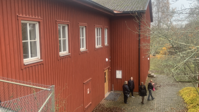 Tre personer står vid entrén till en röd träbyggnad med vitmålade fönster, omgiven av höstträd och nedfallna löv.