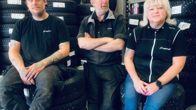 Tre personer sitter på bildäck i ett garage, omgivet av däckstaplar och hyllor med lådor. De bär matchande arbetsuniformer och ser ut att vara personal inom bilindustrin.