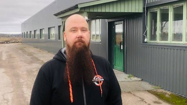 En person med långt skägg står utanför en grå industribyggnad med grönkarmade fönster och en grön dörr en mulen dag.