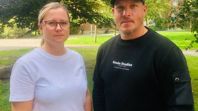 Två personer, den ena i vit t-shirt och glasögon och den andra i svart sweatshirt och keps, står utomhus på en gräsmatta med träd och byggnader i bakgrunden.