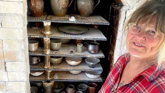 En kvinna i en rutig skjorta ler bredvid en ugn fylld med olika keramiska keramikbitar.