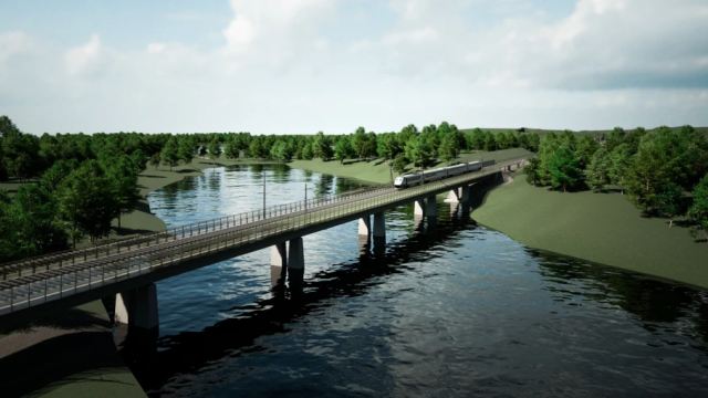 Ett tåg passerar en lång bro över en flod omgiven av gröna träd under en delvis molnig himmel.