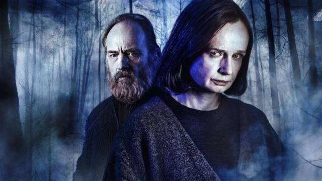 Två allvarliga personer i mörka kläder står rygg mot rygg i en dimmig, blåbelyst skog med kala träd och en bergig bakgrund.