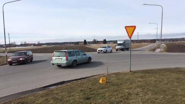 Fyra bilar närmar sig och navigerar i en rondell på landsbygden, med en avkörningsskylt synlig i förgrunden och fält i bakgrunden.