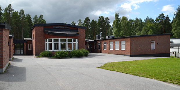 En skolbyggnad i tegel med stora fönster och platt tak, omgiven av träd och en asfalterad innergård under en molnig himmel.