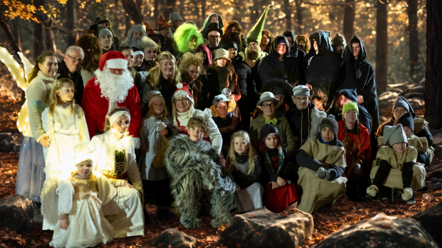 En stor grupp människor klädda i olika fantasi- och djurkostymer poserar tillsammans i en solbelyst skog med höstlöv på marken.