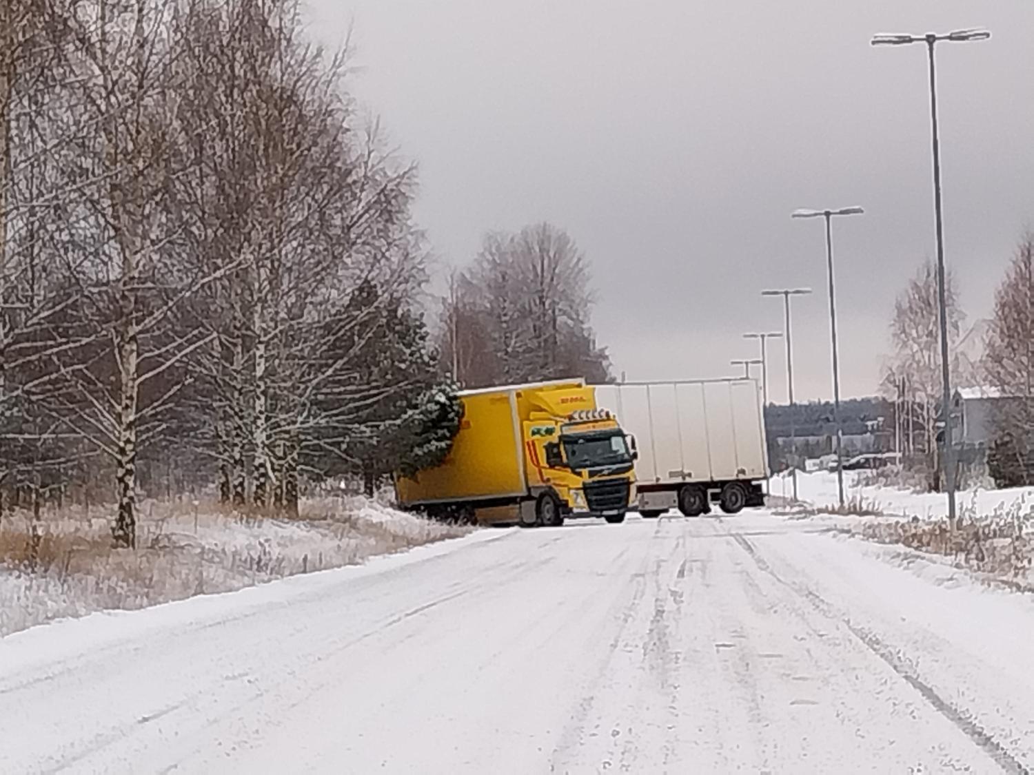 En gul semitrailer med ett vitt släp har kört av vägen på en snöig väg, delvis på vägrenen, och blockerar båda körfälten.