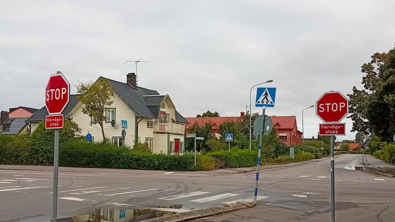 Två stoppskyltar och en övergångsskylt vid en korsning i ett bostadsområde med hus, grönska och molnigt väder.