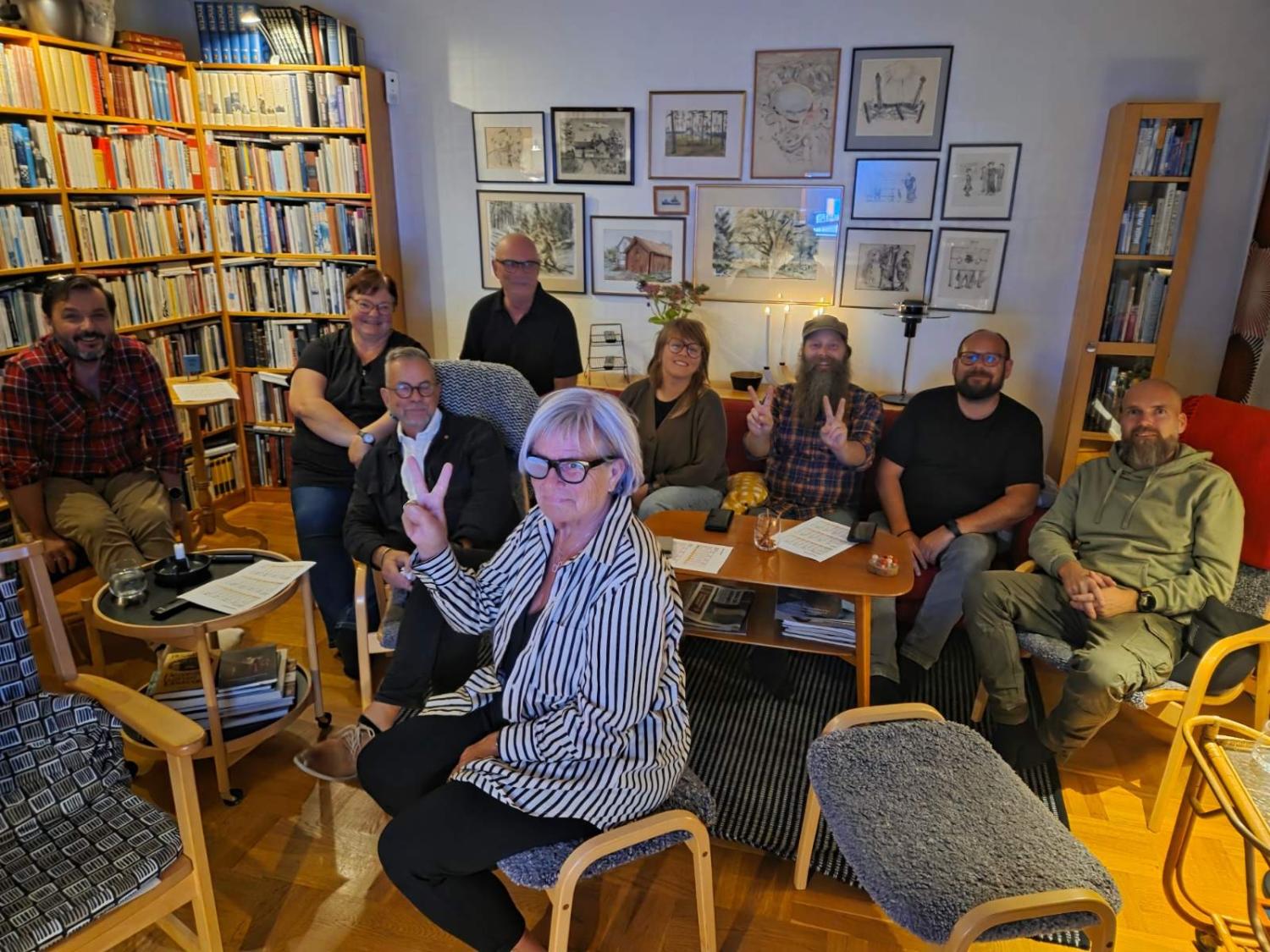 Åtta vuxna sitter tillsammans i ett mysigt vardagsrum med bokhyllor och inramade tavlor på väggarna; en kvinna i förgrunden gör ett fredstecken mot kameran.