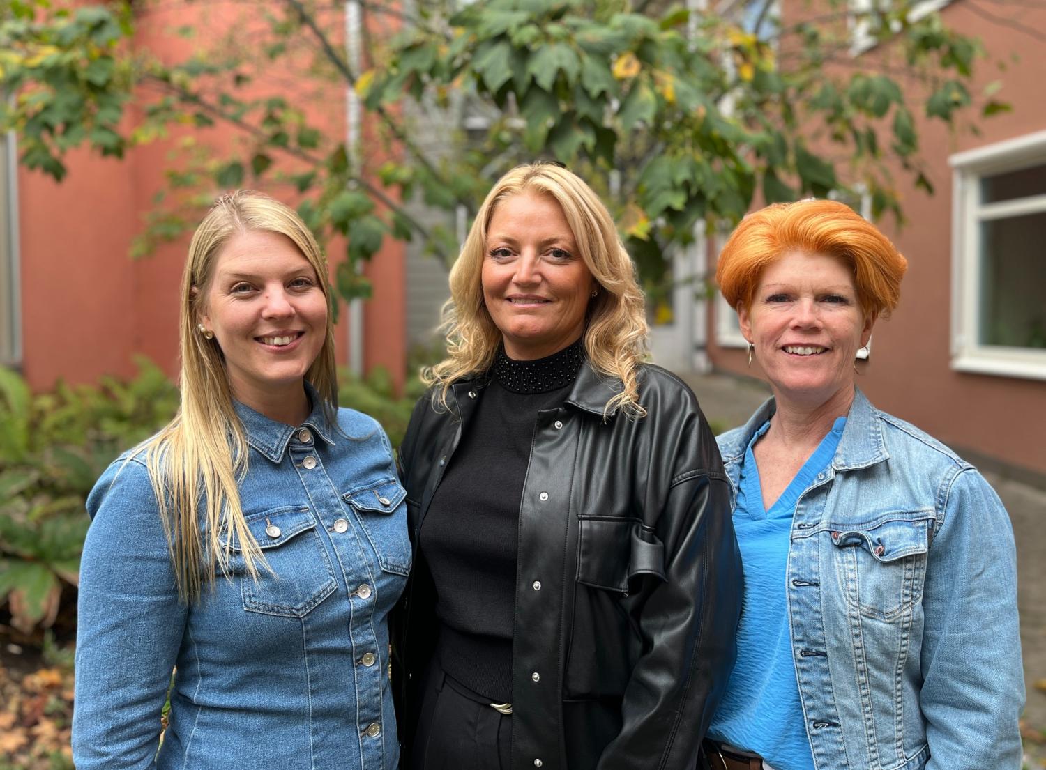 Tre kvinnor står utomhus framför en grönskande miljö och en byggnad och ler mot kameran. Två av dem har på sig jeansjackor, och en har på sig en svart läderjacka.