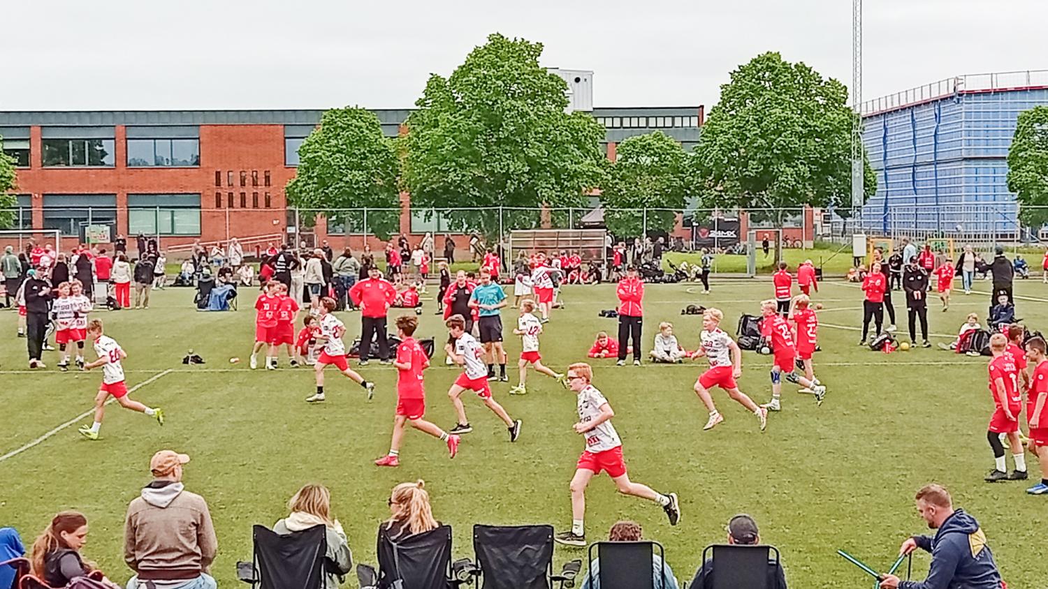 Barn spelar en fotbollsmatch utomhus på en gräsplan, omgivna av tränare, åskådare och människor i rödvita uniformer, med byggnader och träd i bakgrunden.