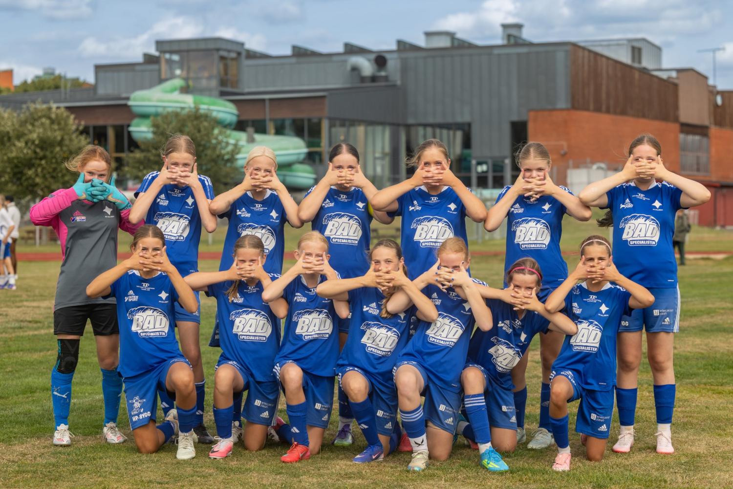 Ett flickfotbollslag i blåa tröjor poserar på en plan, där varje spelare täcker ögonen, öronen eller munnen med händerna. I bakgrunden syns en byggnad och träd.