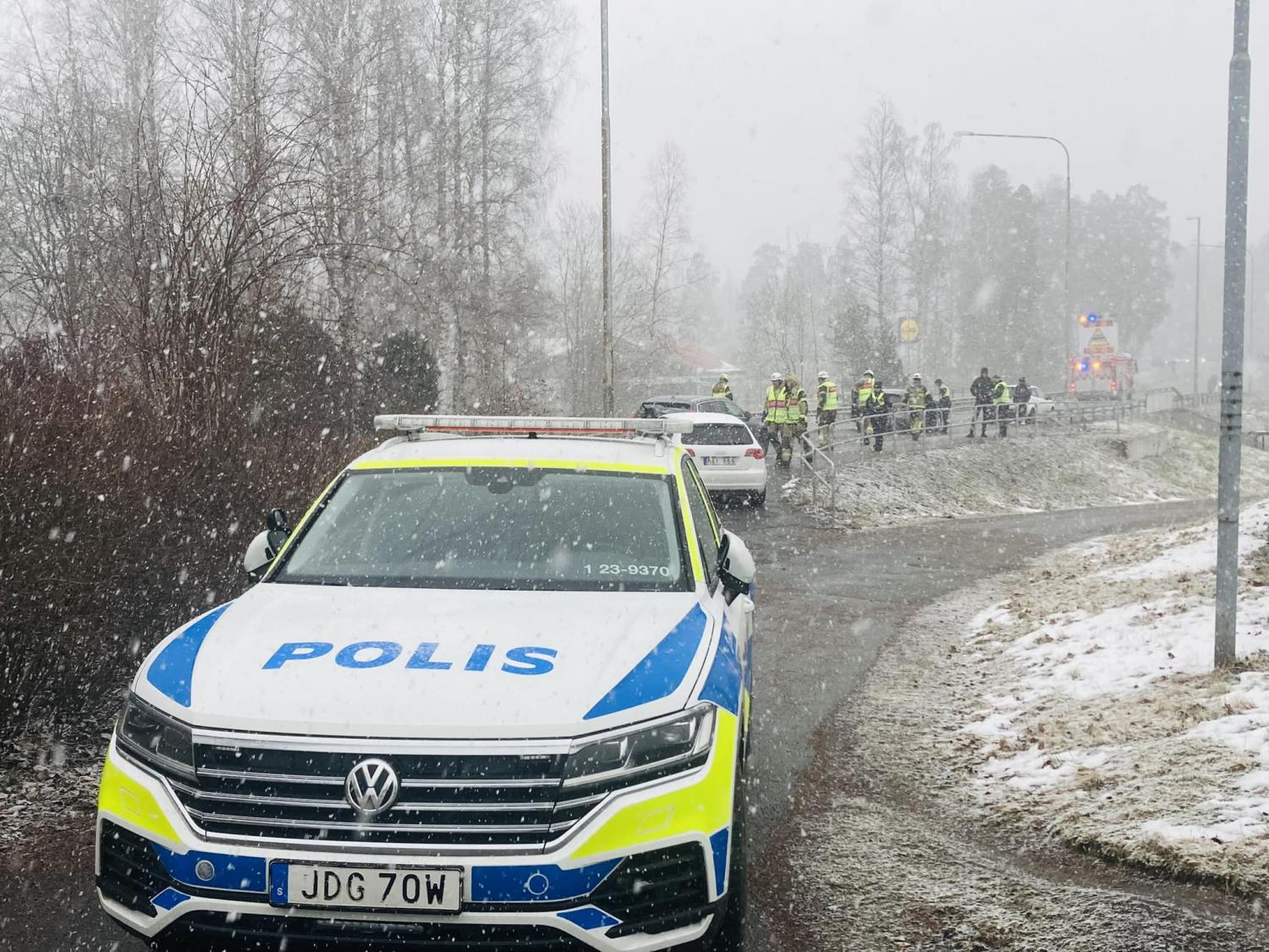En polisbil med tända lampor parkerad vid kanten av en snöig väg, med poliser som står nära en annan incident i bakgrunden.