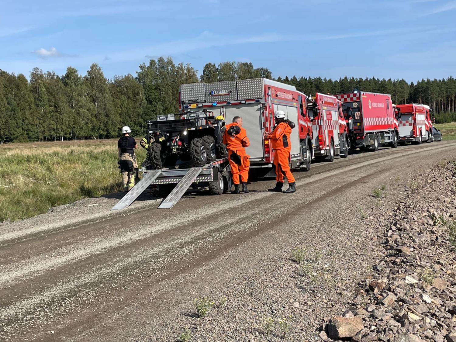Brandmän i orange dräkter lastar av utrustning från en brandbil på en lantlig grusväg, med flera brandbilar på rad och en skog i bakgrunden.