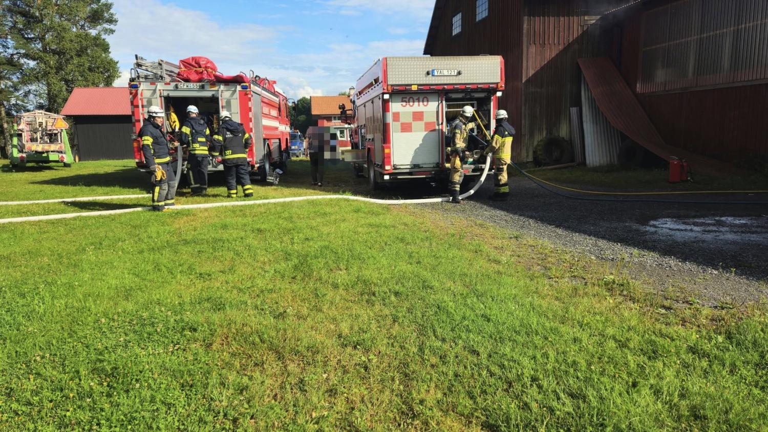Brandmän i full utrustning står nära brandbilar utanför en stor byggnad, med slangar anslutna och vatten på marken.