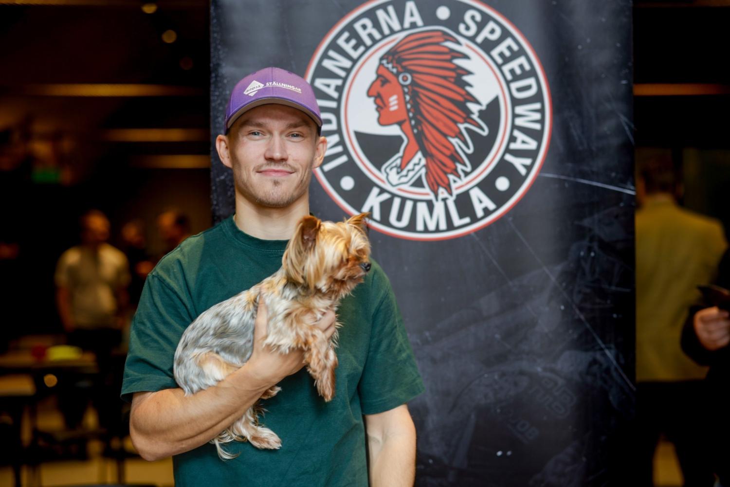En man i grön skjorta och lila keps med en liten hund i famnen står framför en banderoll från Kumla Indianerna Speedway.