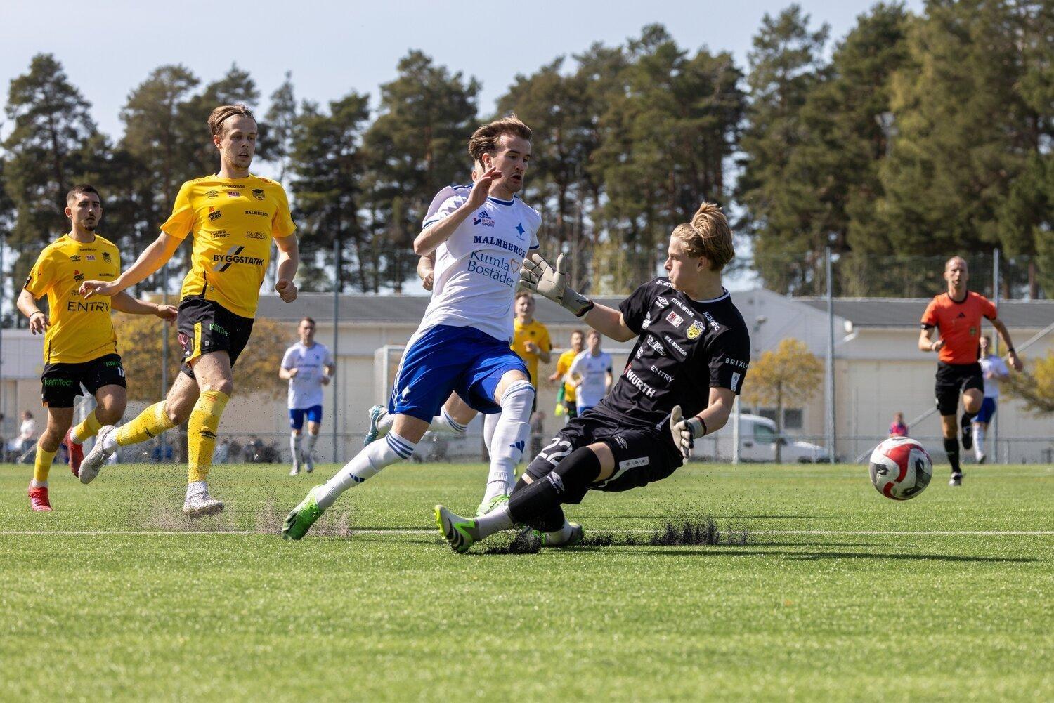En fotbollsspelare i en vit och blå uniform försöker göra mål medan en målvakt i svart dyker för att blockera bollen, med andra spelare och en domare i bakgrunden.