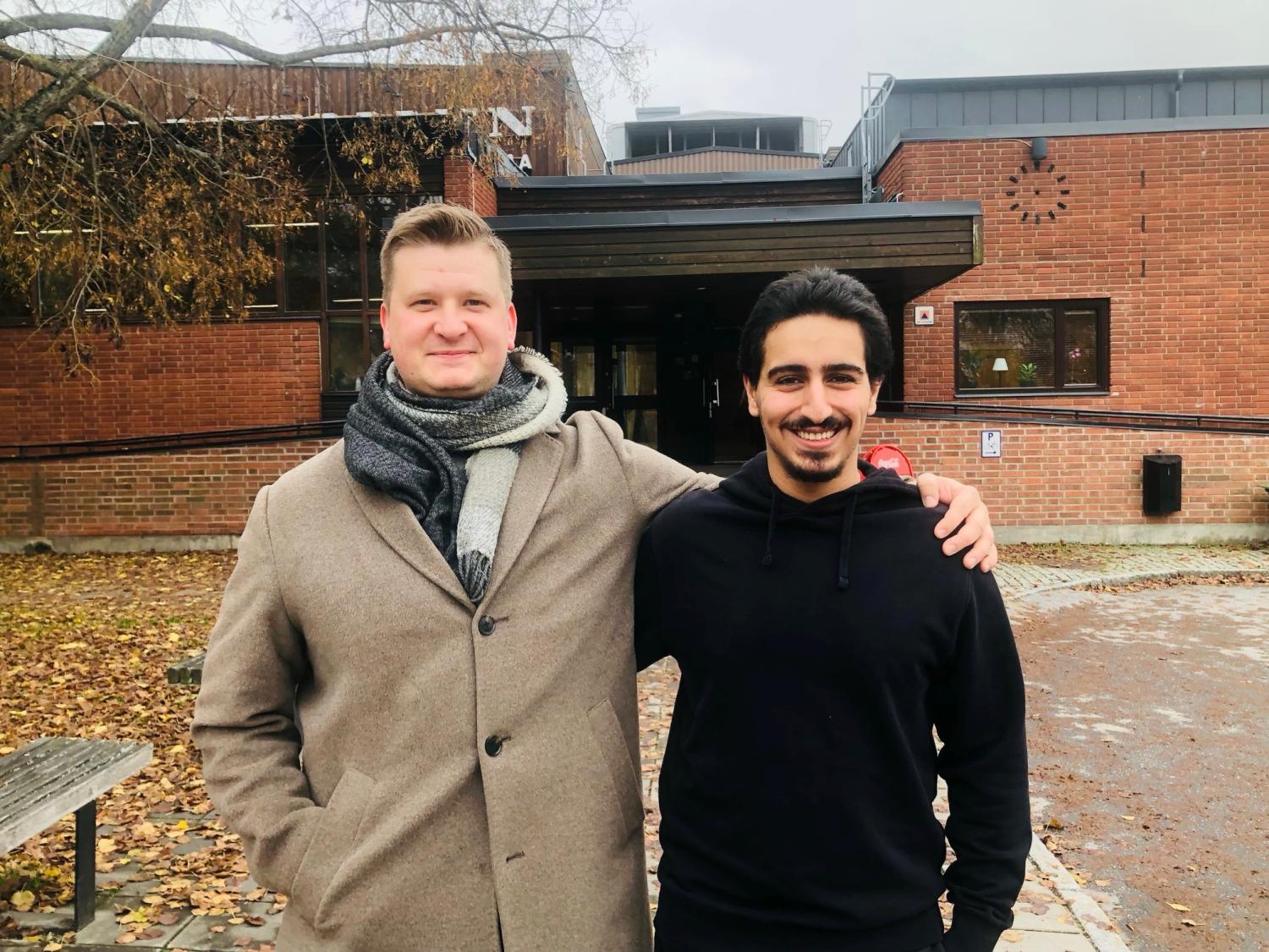 Två män står sida vid sida, den ene i beige kappa och halsduk, den andre i svart huvtröja, och ler framför en tegelbyggnad med höstlöv på marken.