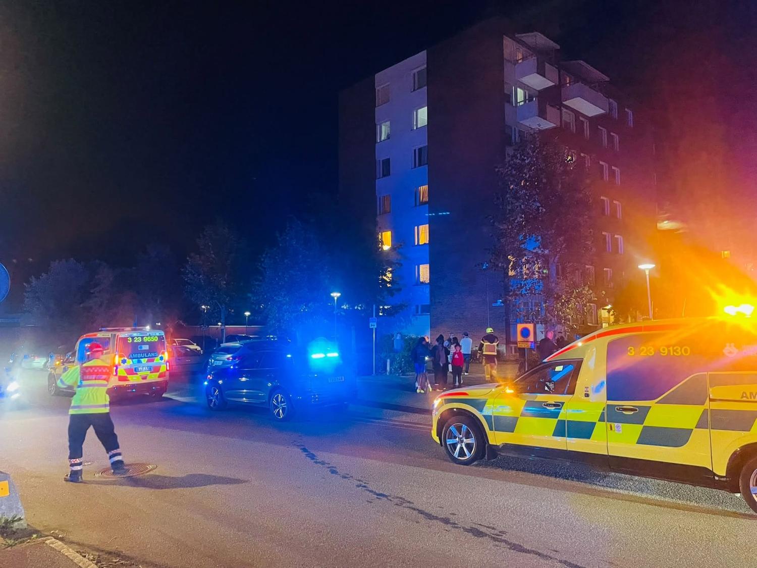 Räddningsfordon med blinkande ljus står parkerade utanför ett flerbostadshus på natten, med människor samlade i närheten och en räddningspersonal som dirigerar trafiken.