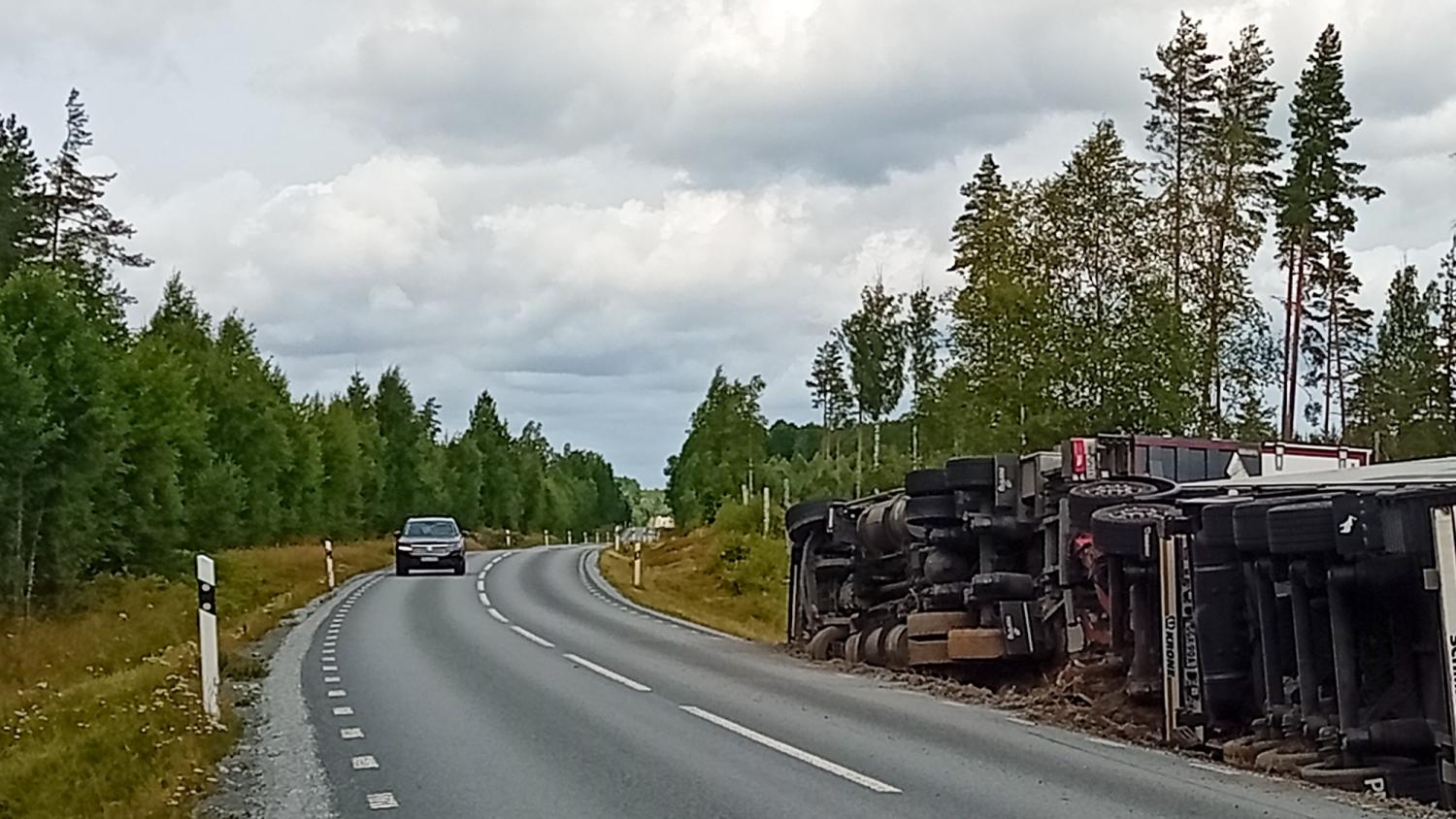 En stor lastbil ligger på sidan vid vägkanten intill en kurvig motorväg, medan en personbil närmar sig från motsatt håll under en molnig himmel.