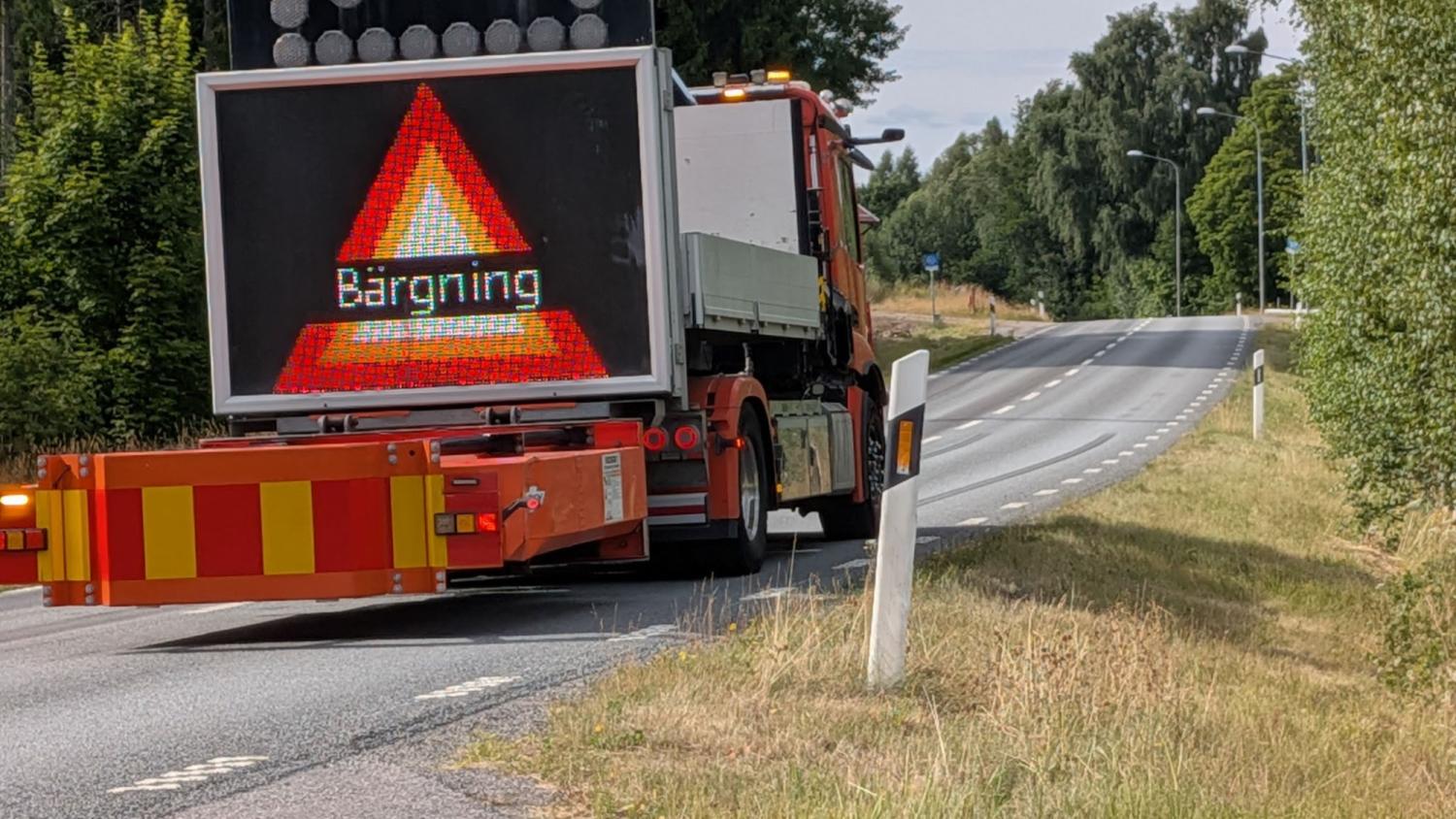 En lastbil med en digital skylt där det står ”Bärgning” och en varningstriangel står parkerad vid sidan av en landsväg med två körfält, omgiven av grönska.