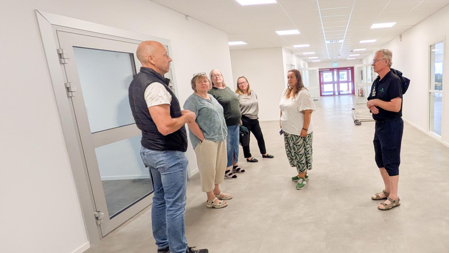 Sex vuxna står i en korridor och pratar med varandra. Korridoren har vita väggar, en glasdörr och synliga taklampor.