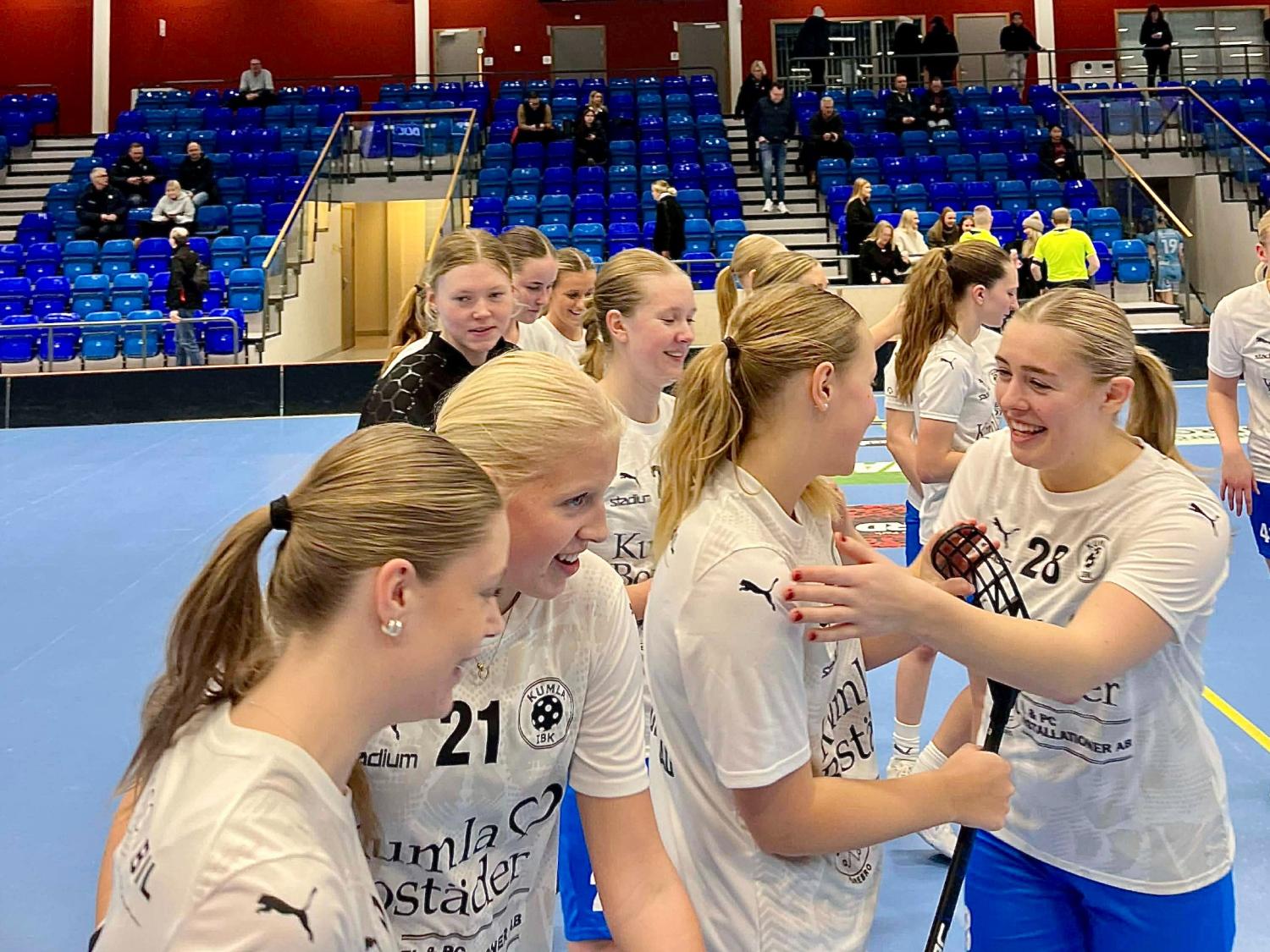 En grupp kvinnliga innebandyspelare i vita tröjor står tillsammans och ler på en inomhusplan, med blå läktare och åskådare i bakgrunden.