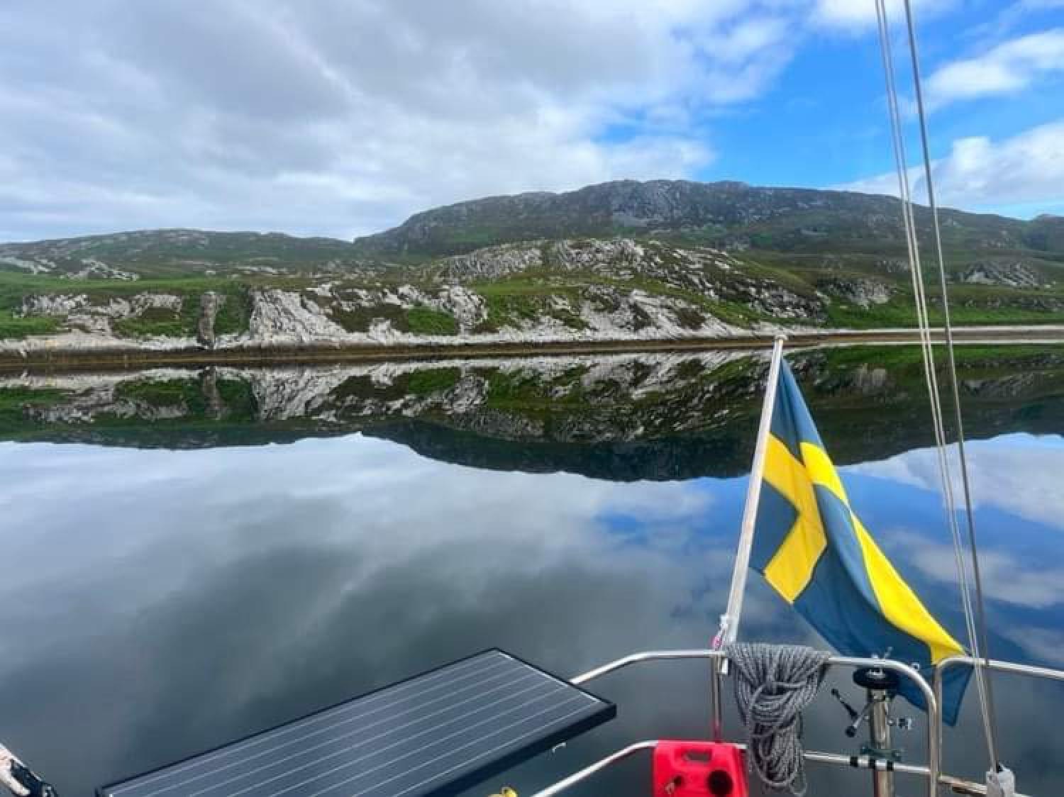 En båt med svensk flagga ligger på lugnt vatten, reflekterande kullar och en halvmolnig himmel i bakgrunden.