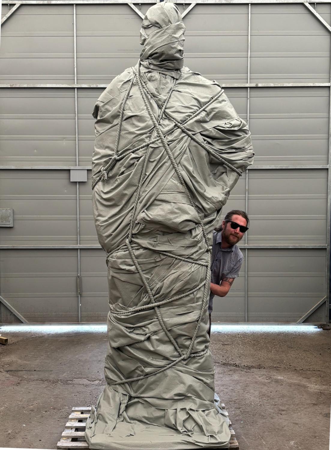 En person med solglasögon står bredvid en stor staty som är tätt insvept i grått tyg, inne i en lagerlokal med en stängd metalldörr.