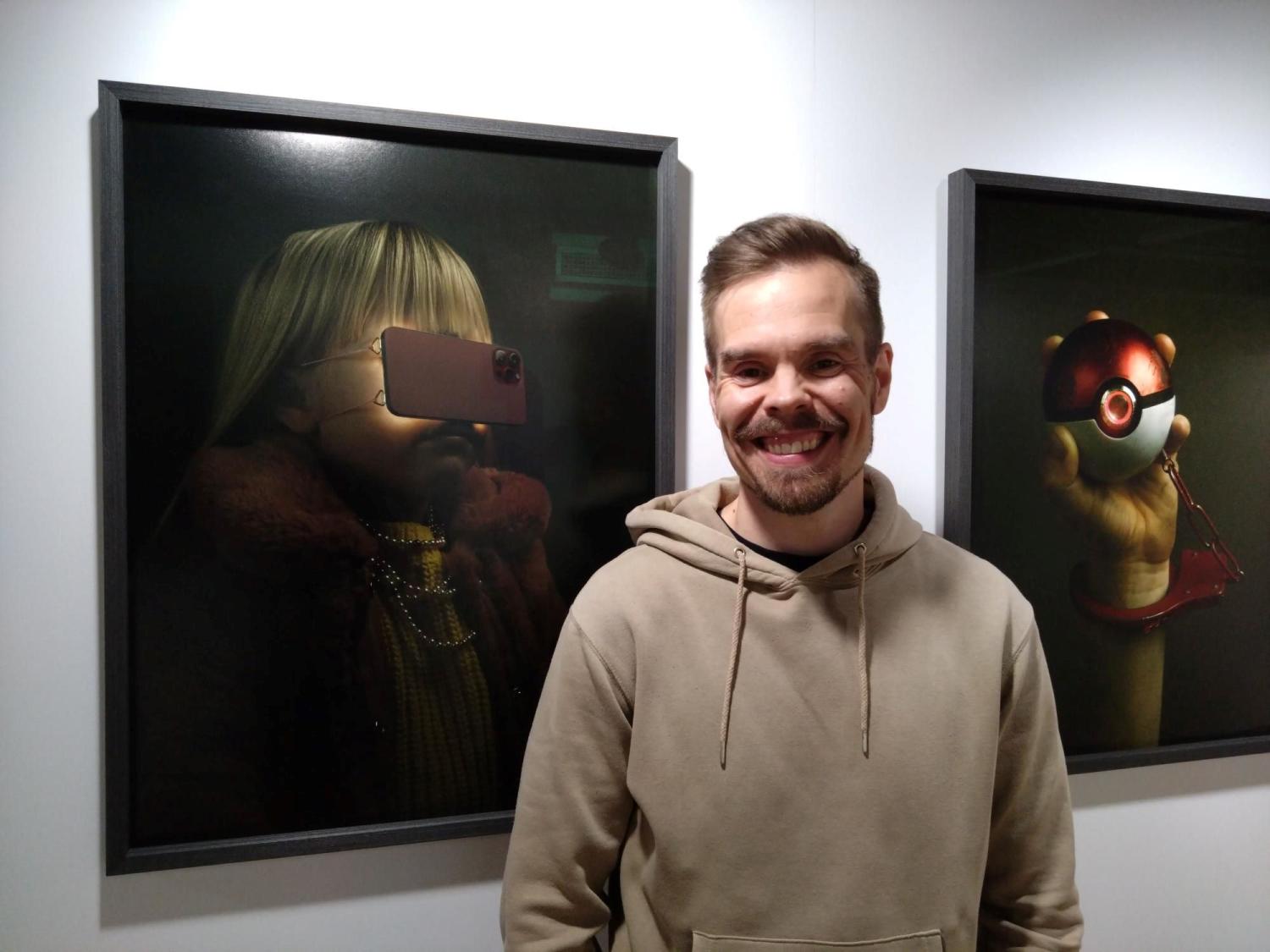 En man i en beige luvtröja står och ler framför inramat konstverk, som inkluderar ett porträtt av en person med virtual reality-glasögon och en bild av en hand som håller ett runt föremål med en röd och vit design.