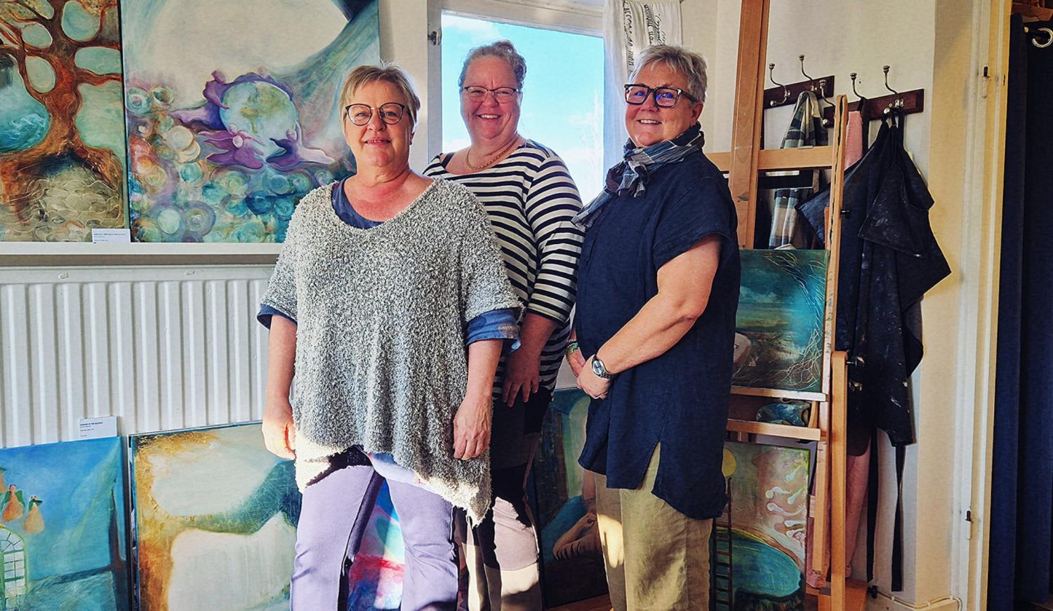 Tre kvinnor står och ler i ett starkt upplyst rum fyllt med färgglada tavlor, bland annat abstrakta verk och naturmotiv, där vissa är utställda på golvet och andra på stafflier och på väggarna.