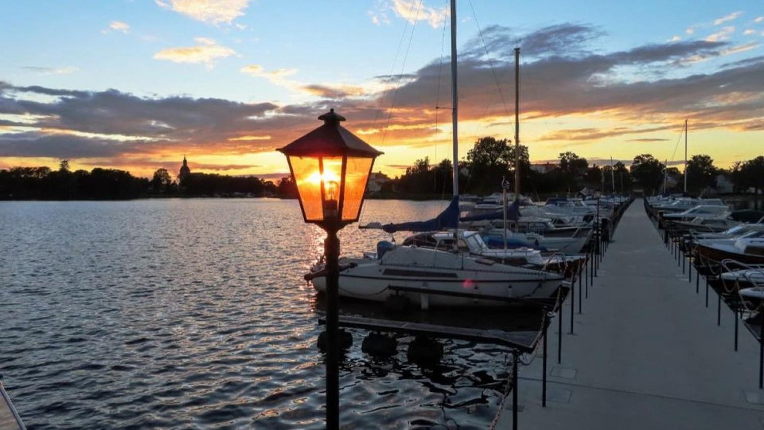 En brygga med båtar på båda sidor vid solnedgången, med en tänd lyktstolpe i förgrunden och lugnt vatten under en delvis molnig himmel.
