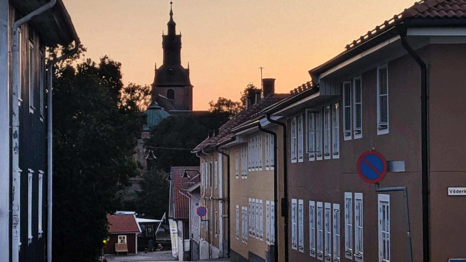 En gata kantad av byggnader vid solnedgången, med ett kyrktorn synligt i bakgrunden och en parkeringsförbudsskylt till höger.