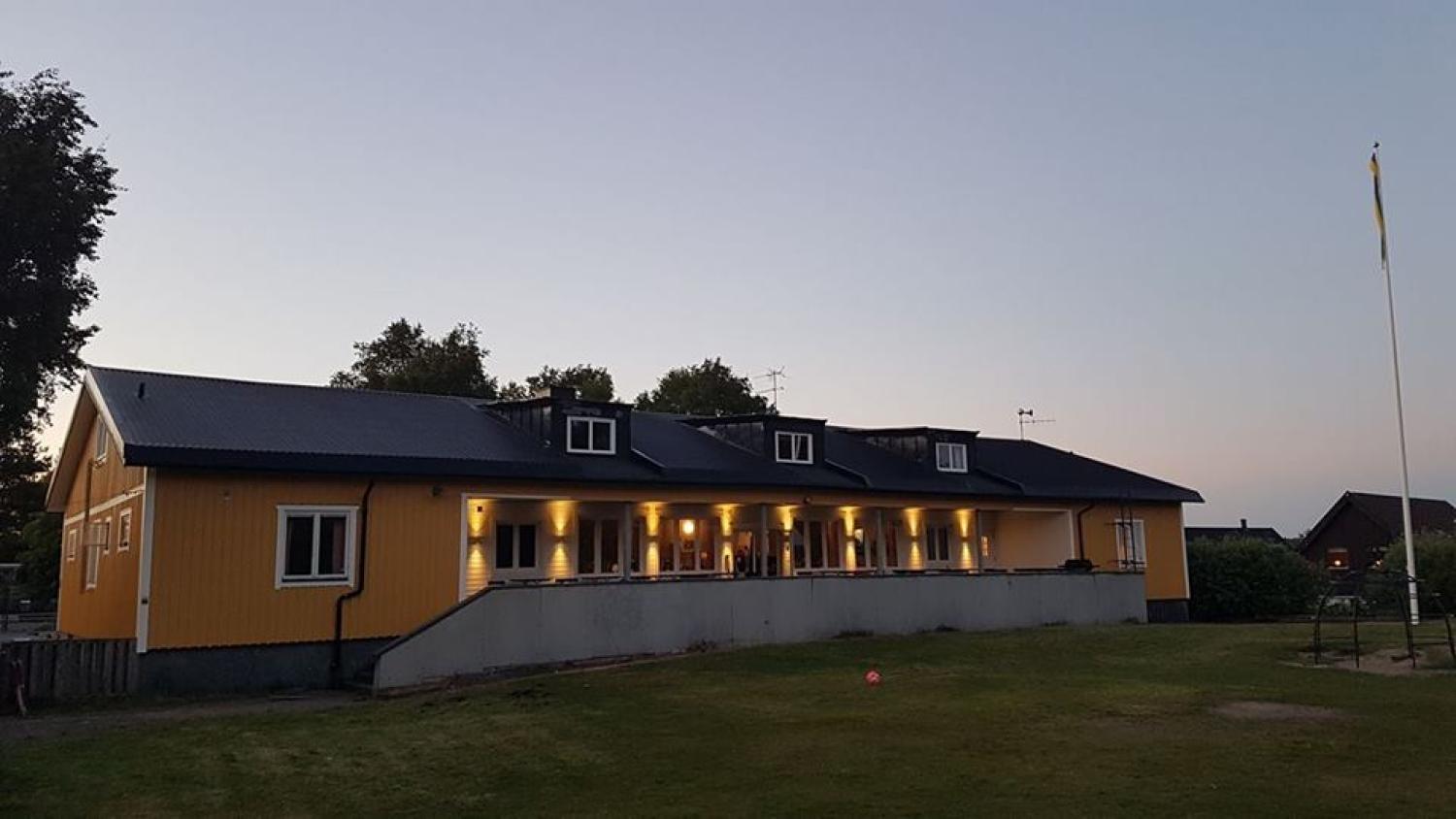 En gul byggnad med mörkt tak, upplyst av ytterbelysning i skymningen, står på en gräsmatta med en flaggstång till höger.