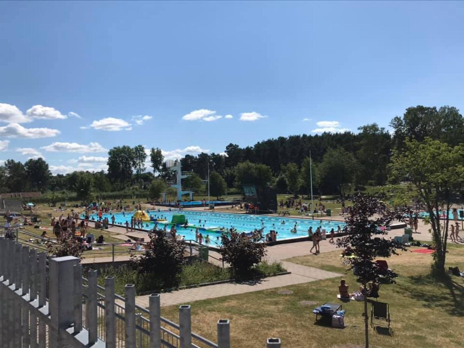 Allmän utomhuspool en solig dag med många människor som simmar, slappar och promenerar runt poolområdet; träd och gräs omger anläggningen.