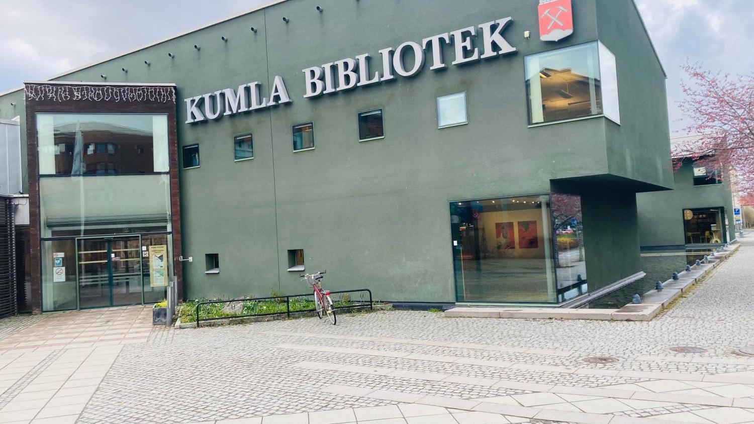 En modern, miljövänlig byggnad med skylten ”Kumla Bibliotek” på fasaden, med stora fönster, en cykel parkerad utanför och en ingång med glasdörrar till vänster.