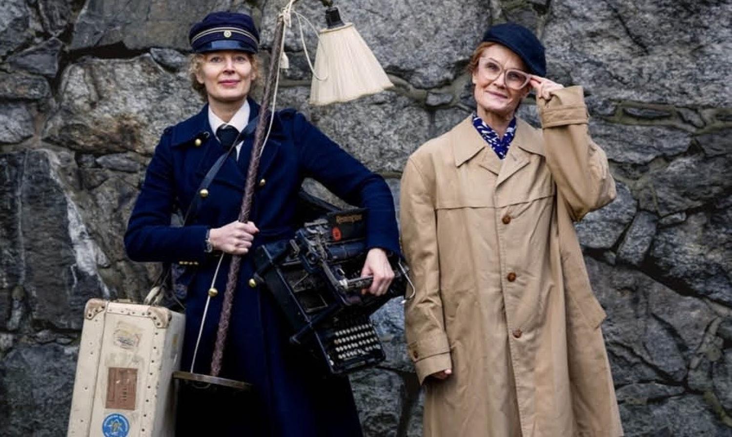 Två kvinnor står framför en stenmur; den ena bär en mörkblå uniform och håller i en skrivmaskin och en lampa, den andra bär glasögon, en beige kappa och en keps och håller i en resväska.