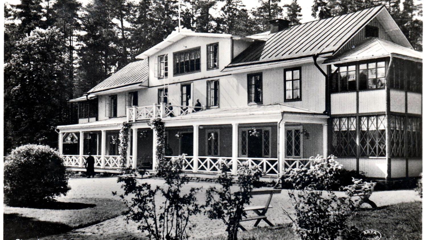 Svartvitt foto av ett stort, tvåvåningshus i trä med en veranda, omgivet av träd och buskar.