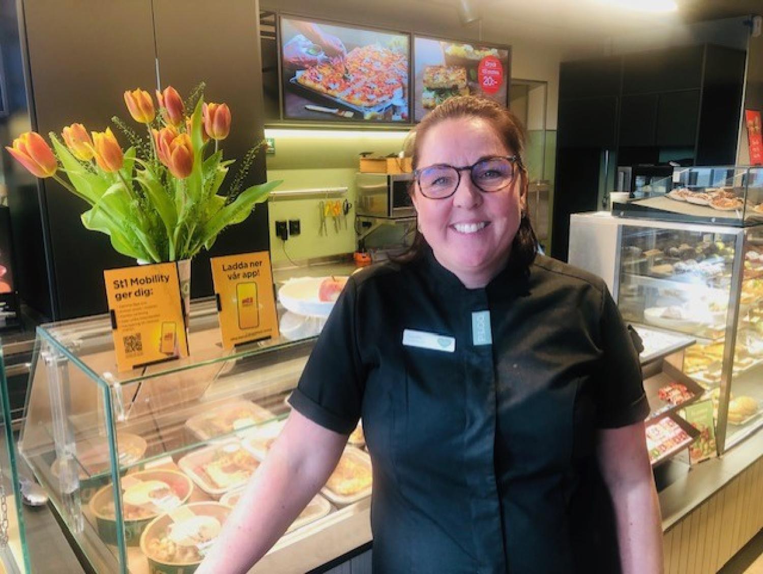 Kvinna i svart uniform står och ler i ett bageri eller en delikatessbutik, bredvid en visning av tulpaner och mat.