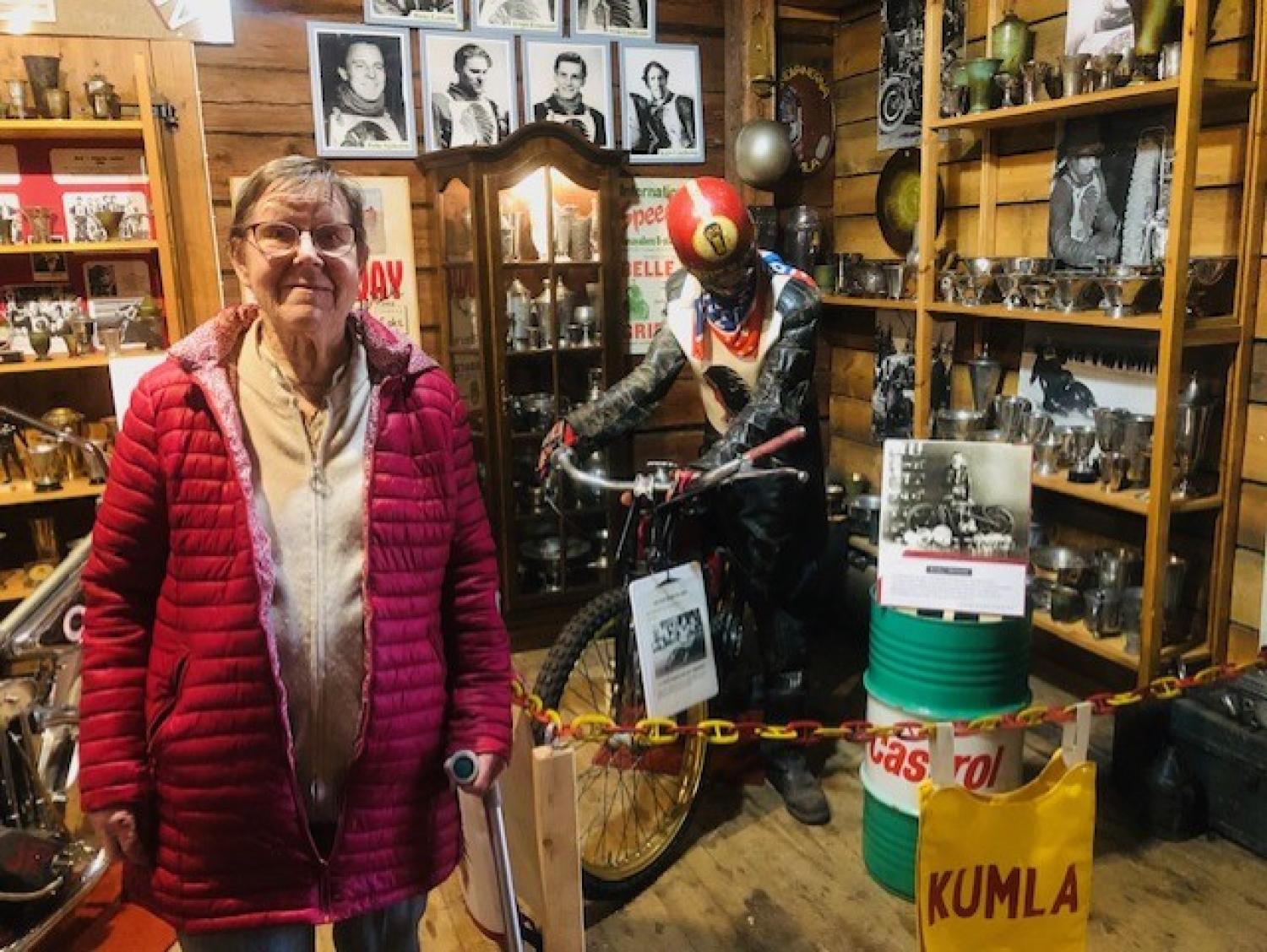 En äldre kvinna i en röd rock står i en vintage motorcykelutställning med minnessaker och foton på väggarna.
